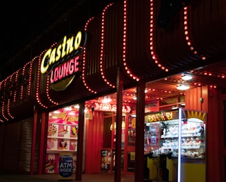 A brightly lit casino lounge entrance adorned with neon lights. The exterior features colorful signage including the words 'Casino' and 'Lounge' in vivid colors, with additional lights framing the building. Inside, a glimpse of a claw machine filled with plush toys and arcade games is visible. The area exudes an inviting yet bustling atmosphere typical of entertainment venues.